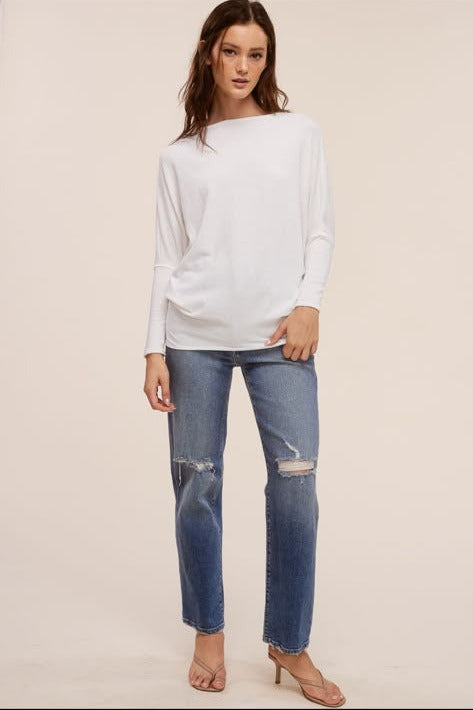 A woman modeling a white long-sleeve boatneck top and blue distressed jeans against a plain beige studio background.