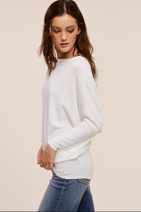 A woman modeling a white long-sleeve knit top with a relaxed, slightly draped fit, paired with blue jeans, set against a solid light beige background.