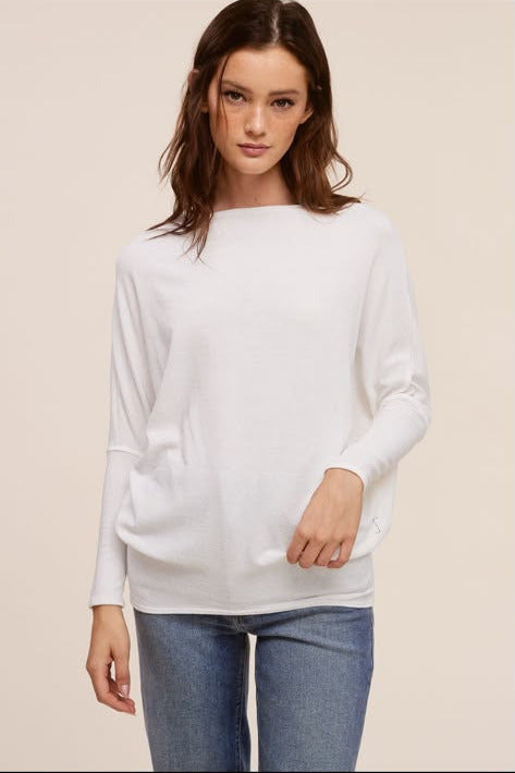 A woman modeling a white long-sleeve knit top with a wide boat neckline, paired with blue jeans, against a solid light beige background.