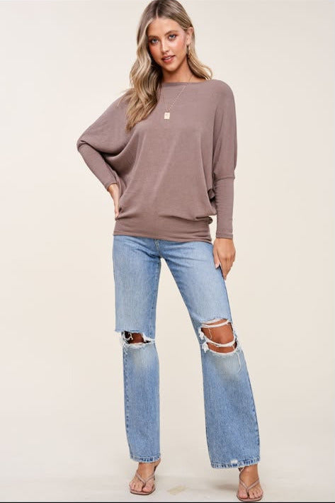 A woman modeling a taupe-brown long-sleeve knit sweater with a small white embroidered logo on the chest, paired with light-wash distressed jeans and flat sandals, against a plain light beige studio background.