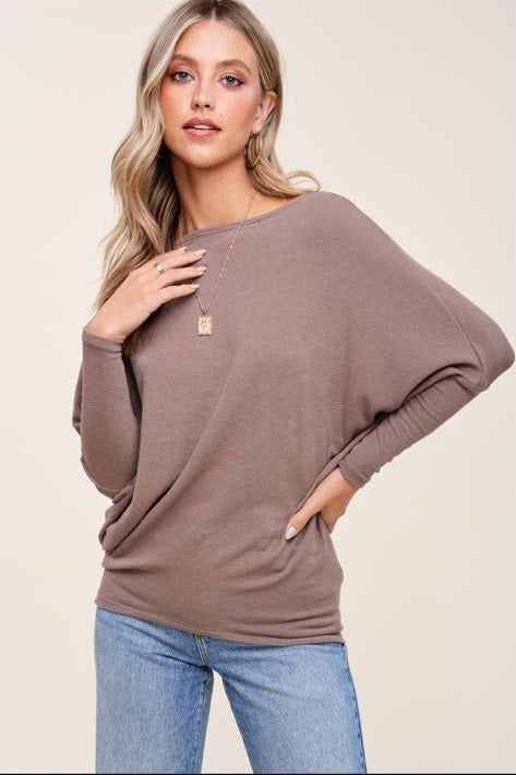 A woman wearing a taupe-brown long-sleeve knit top with a wide boat neckline and ribbed cuffs, paired with light blue jeans, against a solid light beige background.