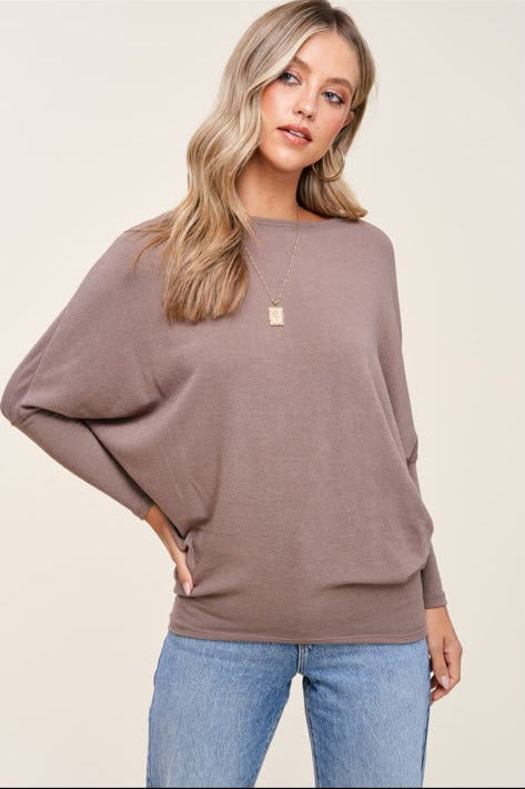 A woman wearing a taupe-brown long-sleeve knit sweater with wide batwing sleeves and light blue jeans, standing against a solid light beige background.