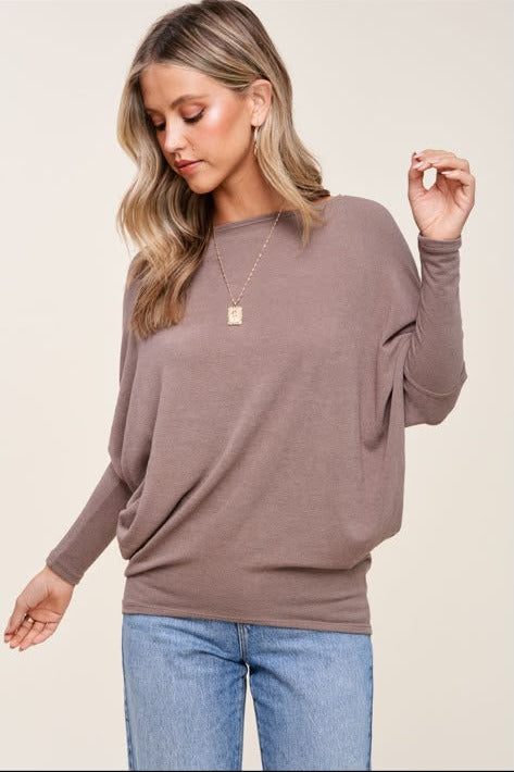 A woman wearing a taupe-brown long-sleeve knit sweater and light blue jeans, standing against a plain light beige background.